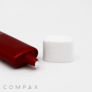 A red 30mm Flat Screw Top tube with a small amount of white cream emerging from its open nozzle lies next to its white cap on a white background. The word "COMPAX" is printed at the bottom left corner of the image.