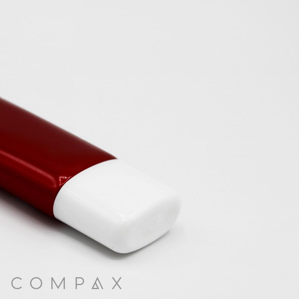 A close-up of a small, red tube with a 30mm flat screw top lies on its side against a white background. The brand name "COMPAX" is visible in the bottom left corner. Only part of the tube is seen, focusing on its cap and a portion of its body.