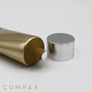 A tube of "40mm Screw Top" skincare product lies on its side with its sleek design glistening, silver cap removed to reveal the sealed opening. The word "COMPAX" is faintly visible at the bottom of the image.