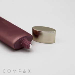 A maroon squeeze tube labeled "35mm Oval Screw Top," with a small amount of cream visible at the nozzle, is lying on its side next to its shiny silver cap. The background is plain white, and the brand name "COMPAX" is seen in the bottom left corner.