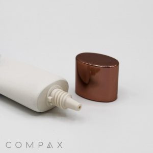 A 25mm Oval Needle Nose is shown with its pointed nozzle tip exposed. The rose gold-colored cap is detached and placed next to the tube. "COMPAX" is printed at the bottom left corner of the image. The background is plain white.