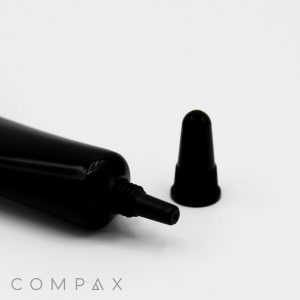 A 16mm Needle Nose tube with a pointed nozzle lies on a white surface, its cap removed and placed nearby. The word "COMPAX" is visible near the bottom left corner.