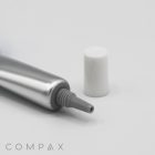 A partially squeezed silver tube with a white cap, positioned on a plain white background. The nozzle of the 13mm Needle Nose tube is visible, and the cap is placed next to it. The word "COMPAX" is located at the bottom left corner of the image.