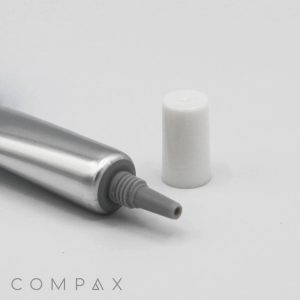 A partially squeezed silver tube with a white cap, positioned on a plain white background. The nozzle of the 13mm Needle Nose tube is visible, and the cap is placed next to it. The word "COMPAX" is located at the bottom left corner of the image.
