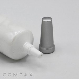 A 22mm Needle Nose squeeze tube with its cap removed is lying on a flat surface. The metallic, cone-shaped cap is placed next to the tube opening. The word "COMPAX" is partially visible at the bottom left corner of the image. The background is plain white.