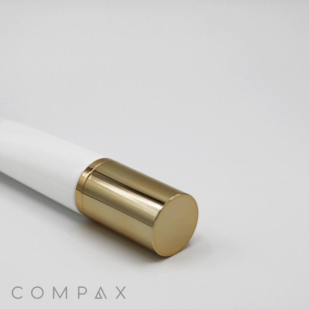 A sleek, cylindrical product named "25mm Airless" with a white body and a golden cap is displayed horizontally on a plain white surface. The brand name "COMPAX" is visible in small letters at the bottom left of the image.