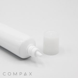 A 25mm Needle Nose tube with a pointed nozzle lies on its side against a white background. The cap, removed from the nozzle, is placed next to the tube. The word "COMPAX" is visible in gray text at the bottom left corner of the image.