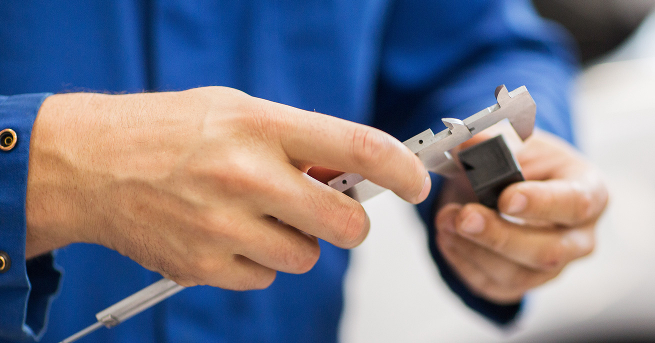Close-up of a person using calipers to measure a small object. The person is wearing a blue shirt, and the focus is on their hands and the measuring tool.