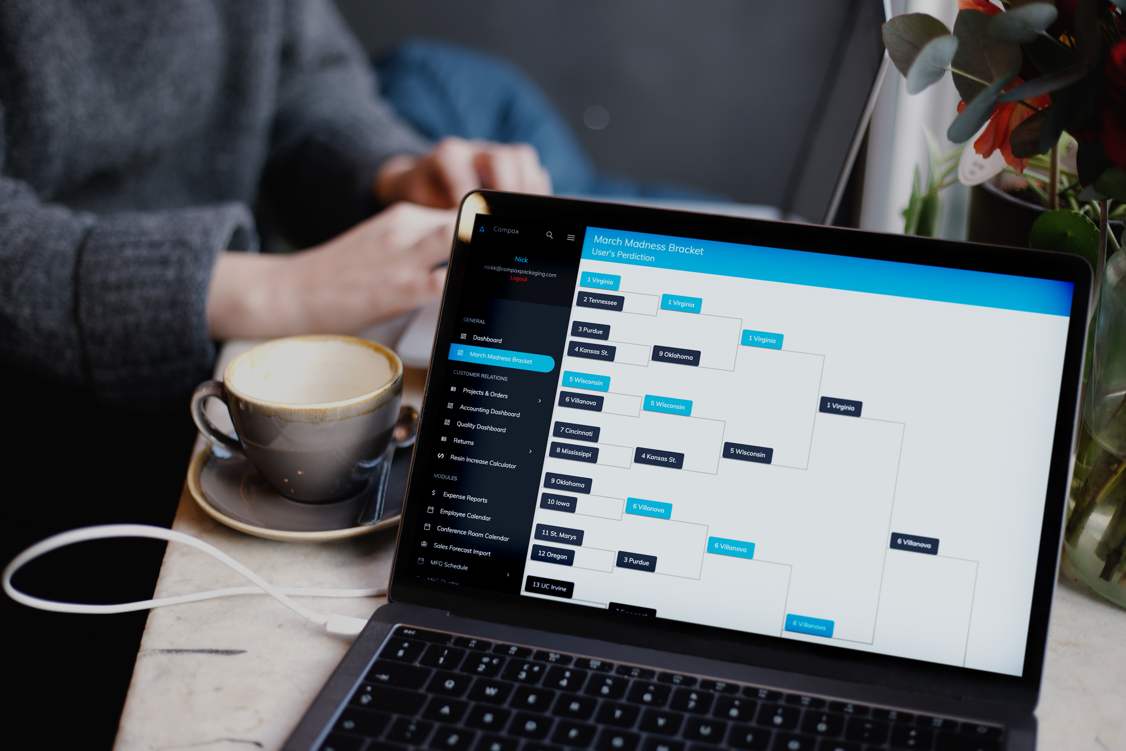 A person sits at a table with a coffee cup, using a laptop. The laptop screen displays a March Madness bracket with team names and matchups. The person appears to be engaged with the screen, likely editing or viewing the bracket. The background is slightly out of focus.