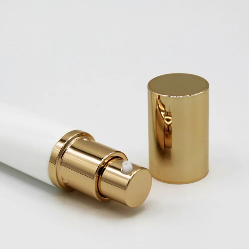 A white airless pump bottle with a gold dispenser pump lid and a gold cap is laying on its side. The cap is removed from the bottle and placed beside it.