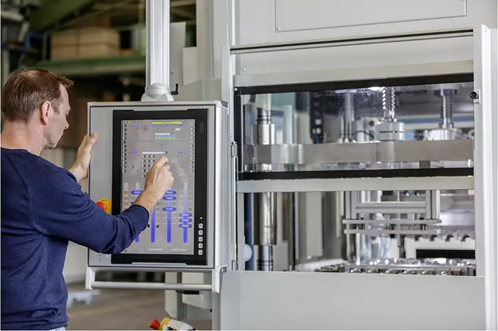 A man operates a large industrial machine using a touchscreen control panel. The machine has visible metallic components and intricate parts, indicating it may be used for manufacturing or processing tasks in a factory or industrial setting.