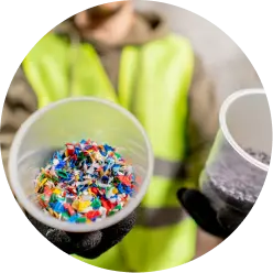 A person wearing a high-visibility vest and gloves holds up two containers. One container is filled with colorful plastic fragments, and the other contains gray plastic shreds. The background is blurred.