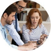 Three individuals are collaboratively working on a project. One person with a beard is listening intently, another with glasses is observing closely, and a third person with long red hair is speaking and gesturing towards a laptop screen. Bookshelves are in the background.
