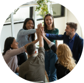 A diverse group of six people in casual and business attire stands in a circle indoors, smiling and giving a collective high-five, conveying a sense of teamwork and celebration. Plants and large windows are visible in the background.