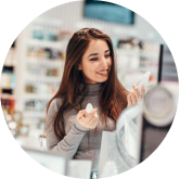 A woman with long brown hair, wearing a gray turtleneck, smiles and examines a skincare product in a store. She holds another item in her left hand, surrounded by shelves stocked with various beauty products.