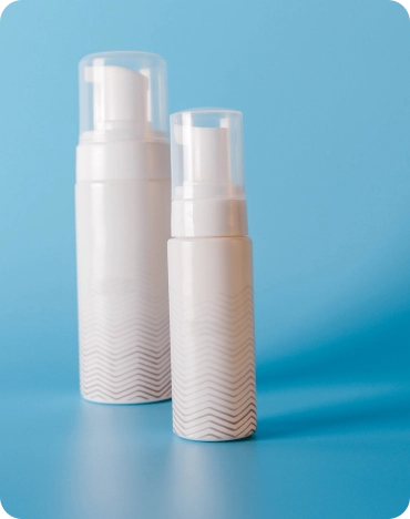 Two white pump bottles with clear caps are positioned against a solid blue background. The bottles feature a subtle wave pattern near the base, with one being significantly taller and slightly larger in diameter than the other.