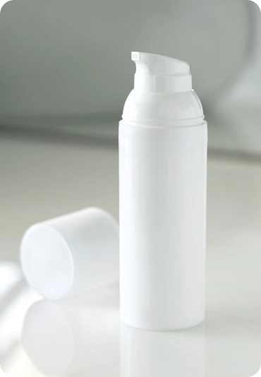 A white plastic bottle with a pump dispenser is standing upright on a light-colored surface. The cap of the bottle is removed and placed next to it. The background is blurred, creating a minimalist and clean appearance.