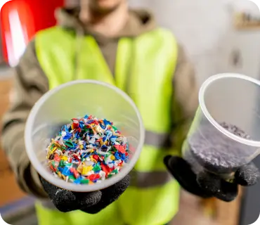 A person wearing a yellow safety vest and black gloves is holding two containers. The container on the left is filled with colorful shredded plastic, while the container on the right holds a dark, granular material.