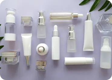 An array of white and clear cosmetic bottles and tubes in various shapes and sizes are arranged neatly on a light-colored surface. Some bottles feature silver accents and droppers. Green leaves are partially visible along the edges of the image.