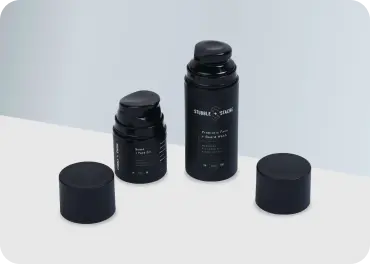Two black airless pump bottles with the caps removed and sitting next to the bottles. The right bottle is a taller than the left.