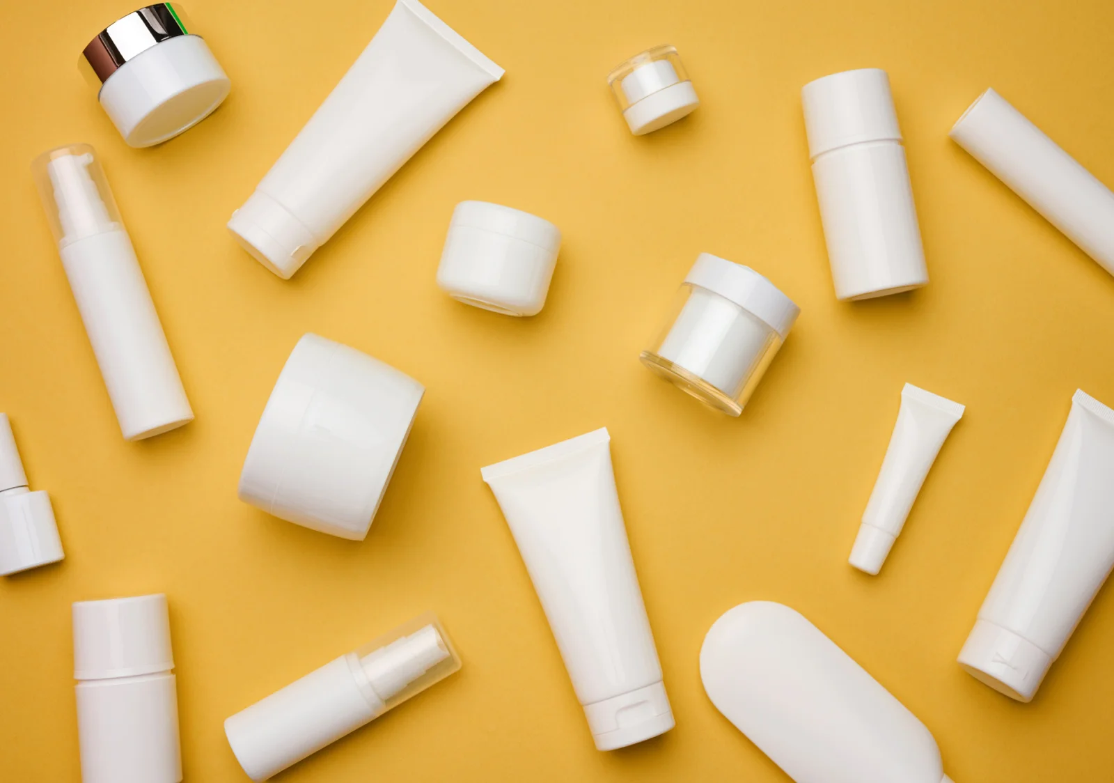 Various white cosmetic containers, including jars, tubes, and pump bottles, are scattered randomly on a yellow background. The containers vary in size and shape, creating an aesthetic arrangement.