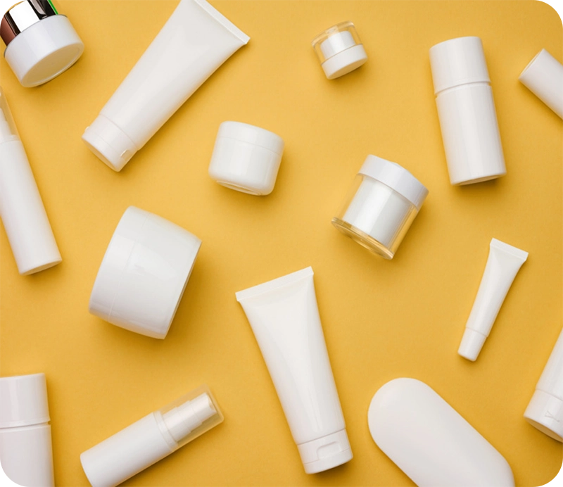 Various white cosmetic containers are scattered on a bright yellow background. The containers include tubes, jars, spray bottles, and pump dispensers, all with a clean, minimalist design.
