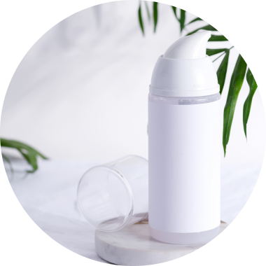 A white foaming dispenser bottle with a blank label is placed on a circular white surface. The clear plastic lid is removed and lies nearby. Green leaves are in the background, with a soft, white fabric backdrop.