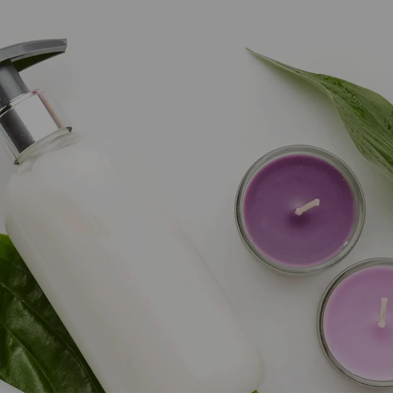 A collection of beauty products on green leaves, including a white bottle with a pump, a beige container with a red pump, and two purple candles in glass holders. The background is light gray.
