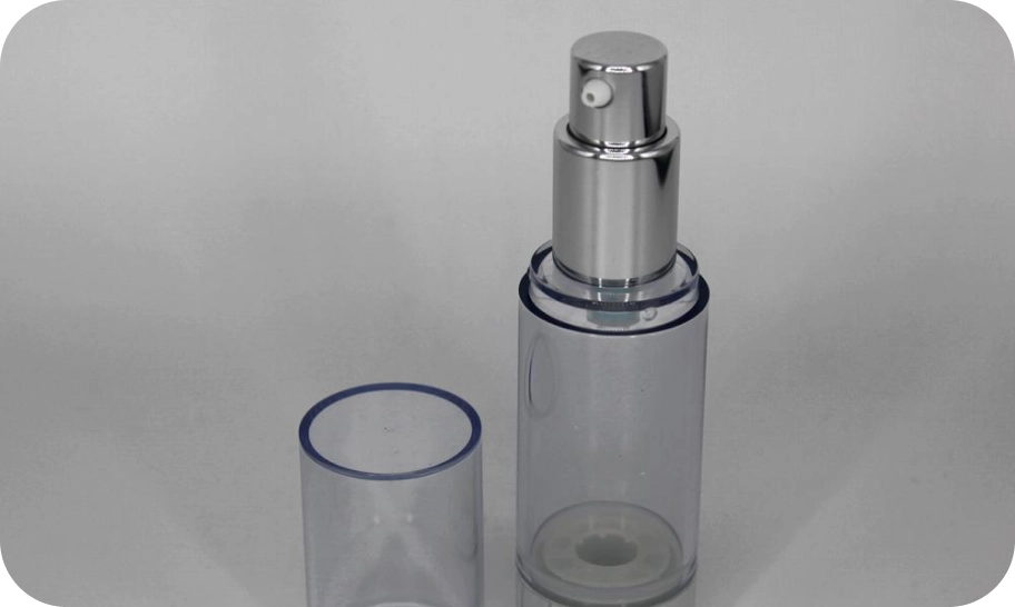 A clear cosmetic spray bottle with a silver pump lies open on a plain background. The cylindrical bottle is accompanied by a matching transparent cap, placed to the side.