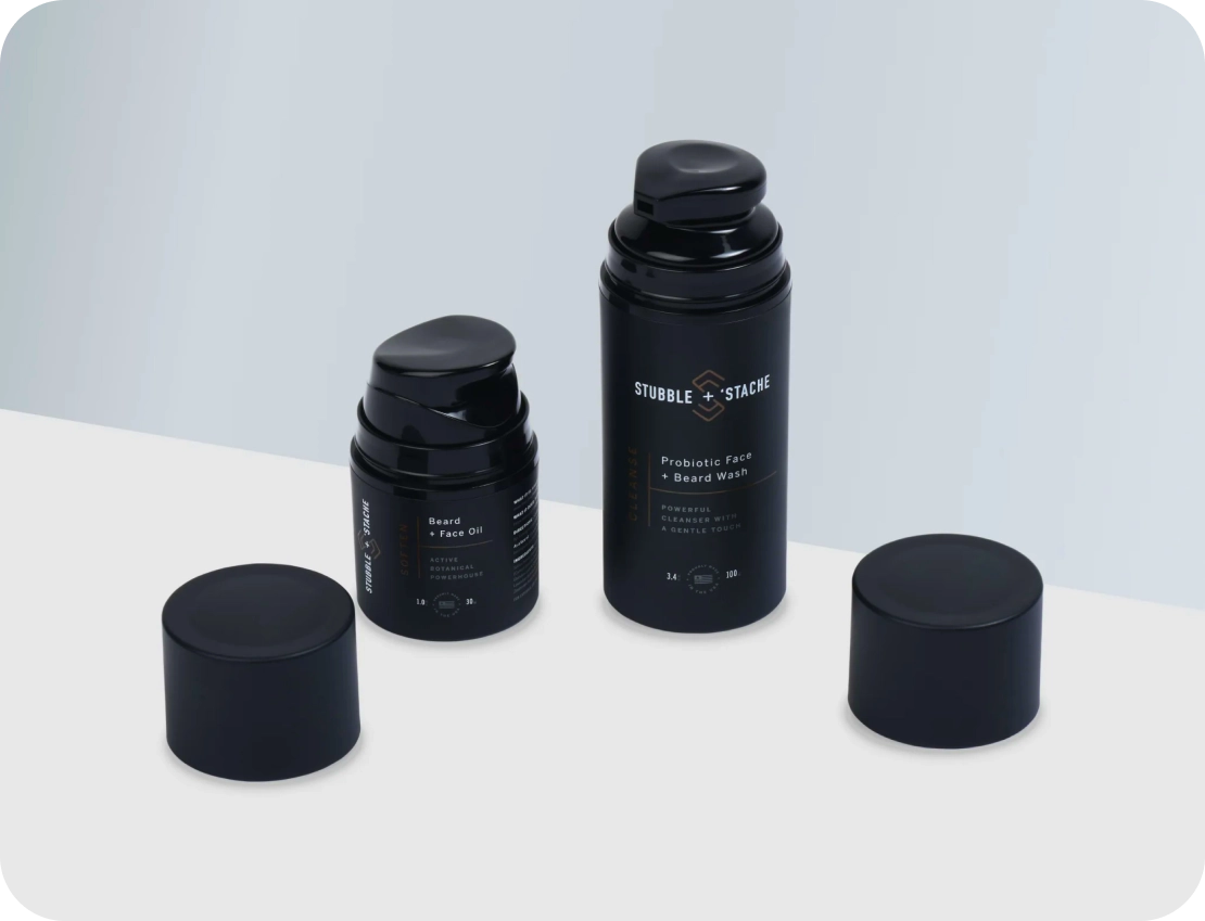 Two black bottles, one larger and labeled Probiotic Face Beard Wash and the other smaller labeled Beard Face Oil, on a white surface with their caps next to them. Both have sleek, minimalist packaging design.