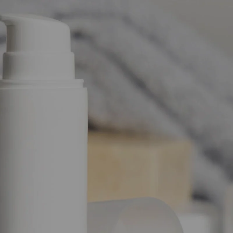 A white pump bottle in focus with a blurred background featuring a folded towel and a beige bar of soap.
