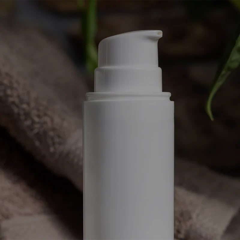White pump bottle in front of a folded brown towel, with potted green plants in the background. The scene is softly lit, creating a cozy and natural ambiance.